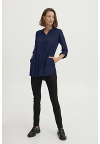 Camicia tunica lunga blu navy con colletto, maniche con bottoni e tasche laterali; abbinata a pantaloni skinny neri e mocassini neri.