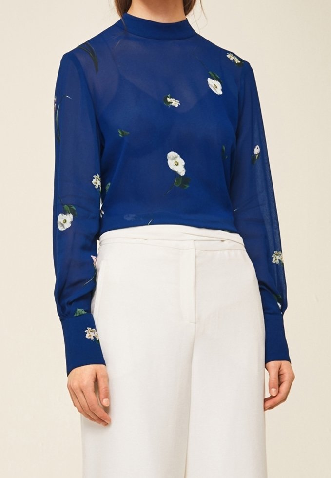 Durchsichtige blaue Bluse mit floralem Muster, hohem Kragen, langen Ärmeln und eng anliegenden Manschetten, kombiniert mit weißen, hoch taillierten Hosen.