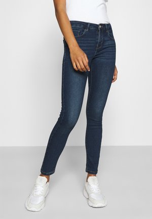 Personne portant un jean skinny bleu foncé, des baskets blanches et un top blanc, debout devant un fond uni gris clair.