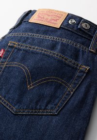 Temno modre kavbojke iz džinsa z oranžnim šivanjem, ki vključujejo usnjen znamčni obliž in značilno obliko zadnjega žepa. Vidna je kovinska sponka za zapiranje.