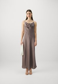 Ilga, be rankovių suknelė iš blizgaus mauve satino audinio, su grotelėmis iškirpte. Modelis laiko mažą baltą rankinę. Švelni ir lygi tekstūra.