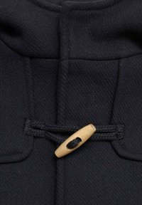 Närbild av en svart texturerad tygkappa med en träspänneknapp och en flätad snoddslut