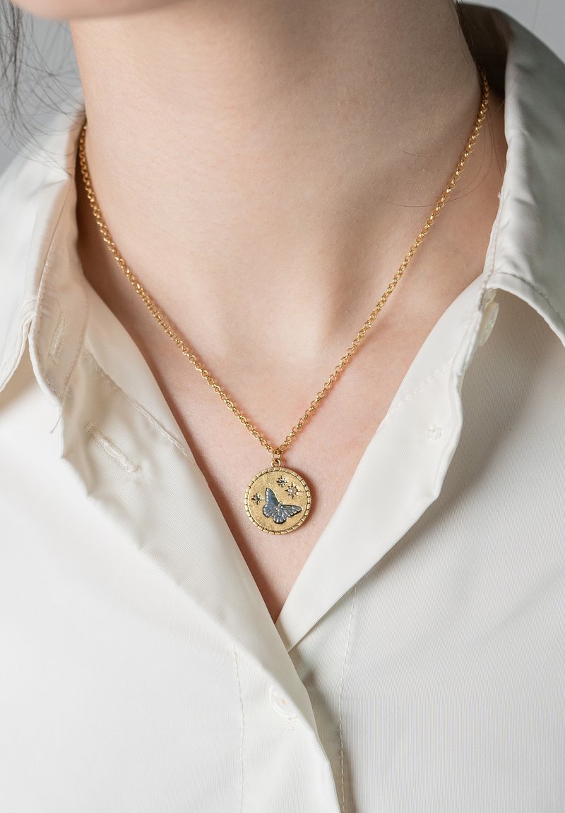 Kullast kett-kaelakee, millel on ümmargune ripats sinise liblikaga ja kolm väikest tähelist disaini, kantud valge kraega särgi peal.