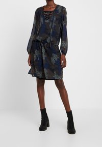 Dlouhé šaty v tmavě modré barvě s květinovým vzorem ve světle modré a hnědé, s šněrovacím výstřihem a elastickým pasem, zkombinované s černými botami.