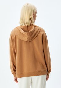 Sudadera marrón con una textura suave, corte relajado y una capucha amplia. Puños y dobladillo acanalados, con un diseño minimalista.
