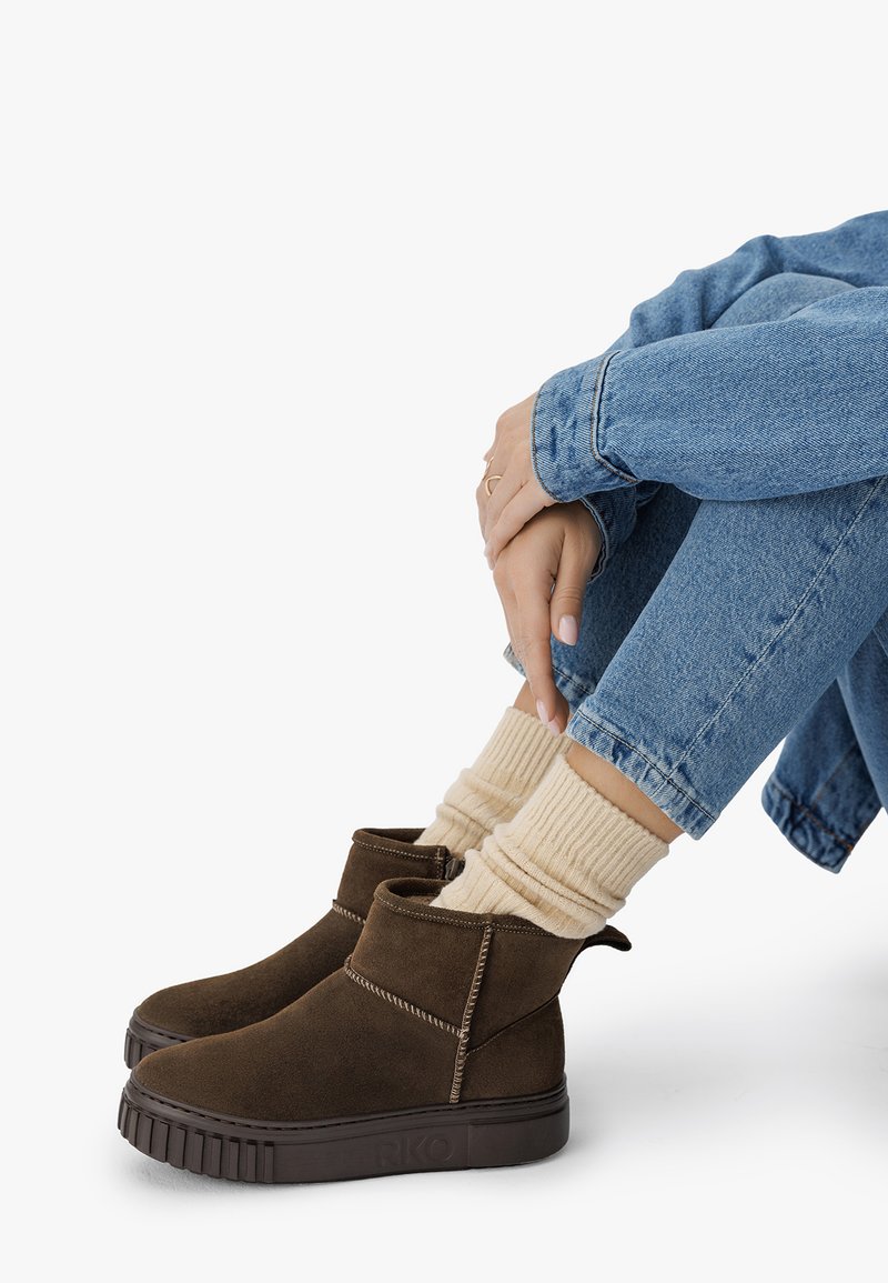 Person som bär mörkbruna mockastövlar, krämfärgade ribbade strumpor och blå jeans, sitter med händerna vilande på korslagda knän.