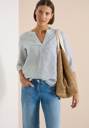 Nainen, jolla on vaaleanharmaa raidallinen pusero ja siniset farkut, kantaa suurta kudottua vaaleanruskeaa olkalaukkua.