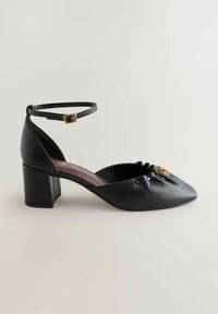 Chaussure en cuir noir à petit talon carré, avec bride de cheville et boucle dorée, présentant un devant froncé et un détail décoratif doré.