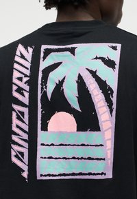Crna majica s poleđinom na kojoj se nalazi pastelni grafički prikaz palme, sunca, valova i okomiti tekst "SANTA CRUZ" na lijevoj strani.