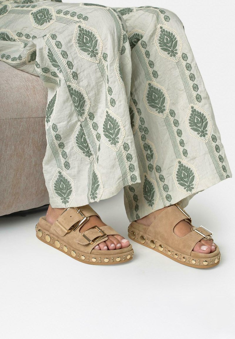 Personne portant des sandales slip-on beiges à clous et un pantalon à jambes larges avec une broderie florale verte, assise sur un canapé marron clair.