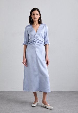 Himmelblaues, gestreiftes, langärmliges Kleid mit V-Ausschnitt, gerafftem Bund und gerader Silhouette. Kombiniert mit hellen, flachen Schuhen.