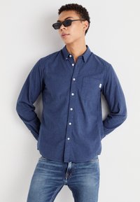 Camisa de botones azul marino en una tela texturizada, con un bolsillo en el pecho y botones blancos, combinada con jeans de mezclilla y gafas de sol oscuras.