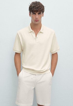 Polo de couleur crème avec un tricot texturé, manches courtes et col traditionnel. Porté avec un short de couleur claire.