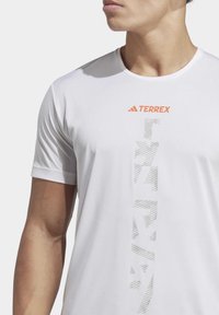 Camiseta deportiva blanca con cuello redondo, mangas cortas y un patrón geométrico gris con el logotipo naranja "TERREX" en el pecho.