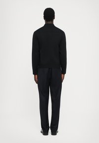 Černá halenka s dlouhými rukávy, límečkem a rovním spodním lemem, v kombinaci s tmavými kalhotami. Hladká látka s minimalistickým designem.