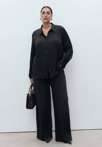Camisa negra de manga larga con botones y textura suave, combinada con pantalones anchos negros y un pequeño bolso marrón.