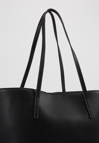 Bolso tote de cuero negro texturizado con tirantes delgados sobre un fondo claro y liso.