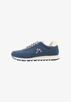 Zapatilla deportiva azul y beige con cordones morados, suela texturizada y un logo de pájaro en el lateral, vista desde el lado exterior.