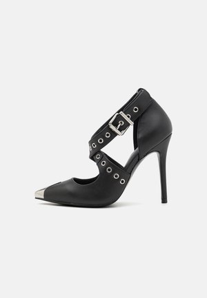 Chaussure à talon haut noire avec bout en métal pointu, brides croisées avec boucle et détails en œillets argentés sur fond clair uni.