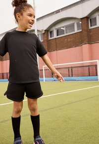 Maglietta nera a maniche corte e pantaloncini, abbinati a calze nere fino al ginocchio e scarpe sportive, su un campo di erba artificiale verde.