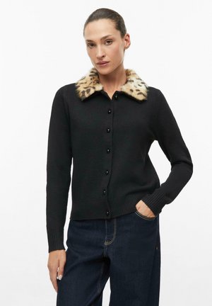 Frau trägt eine schwarze, geknöpfte Strickjacke mit Leopardenfellkragen und dunkelblaue Jeans, steht mit einer Hand in der Tasche.