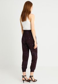 Pantalon noir à motifs floraux avec une taille élastique se resserrant aux chevilles. Le mannequin porte des talons noirs à lanières et un débardeur blanc à côtes.