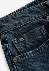 Tamne traperice od tamnog denima koje imaju prednji džep s crnim tekstilnim oznakom i metalnim dugmetom, kontrastne šavove i teksturiranu površinu.