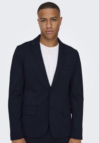 Blazer navy en una tela suave, de una sola fila de botones con dos botones. Cuenta con solapas con muesca, un bolsillo en el pecho y dos bolsillos frontales.