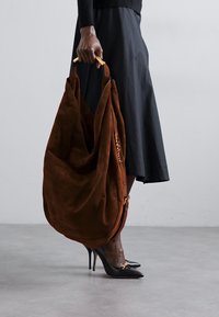Grand sac en suède marron au design décontracté. Présente des accents en métal doré. Porté avec une jupe noire mi-longue et des talons hauts noirs.
