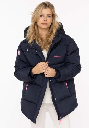 Blonde Frau trägt eine marineblaue Steppjacke mit pinkfarbenen Reißverschlüssen und Kapuze und schließt den vorderen Reißverschluss der Jacke.