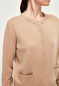 Cardigan beige avec un col rond, fermeture à cinq boutons et deux poches avant. Fabriqué en matériau doux et texturé avec une finition lisse.