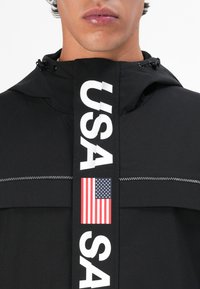 Sort jakke med hætte, hvid "USA" skrift og et amerikansk flag patch. Stoffet har en struktureret overflade med synlige sømme.