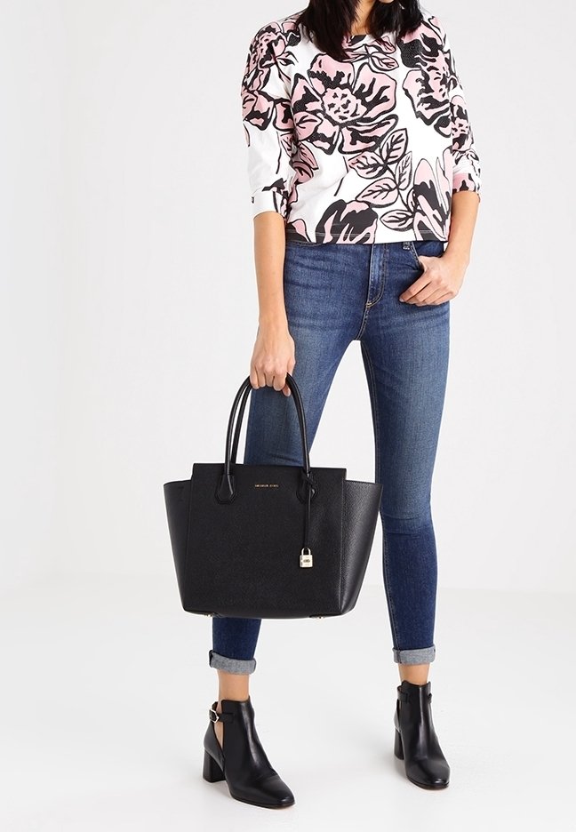 Sac fourre-tout en cuir noir au design structuré, avec des poignées courtes et une étiquette en métal. Porté avec un haut à motifs floraux et un jean en denim foncé.