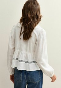 Vrouw met lang bruin haar, gekleed in een witte blouse met lange mouwen met kantdetails en een blauwe spijkerbroek, met haar rug naar de camera gekeerd tegen een effen muur.