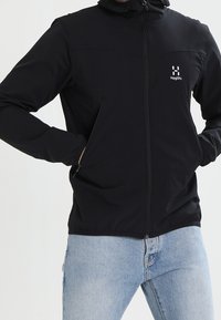 Veste noire à fermeture éclair avec capuche, fabriquée en tissu doux, avec un panneau cousu sur la poitrine et des poches latérales. Logo sur le côté gauche.