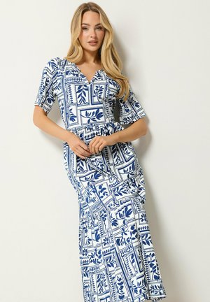 Lyshåret kvinde iført en hvid midi-kjole med et blåt botanisk og geometrisk mønster, korte ærmer og en V-udskæring, poserende foran en hvid væg.