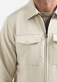 Lichtbeige jas met een ritssluiting, twee borstzakken en een puntige kraag. Gemaakt van een gladde stof met een subtiele textuur.