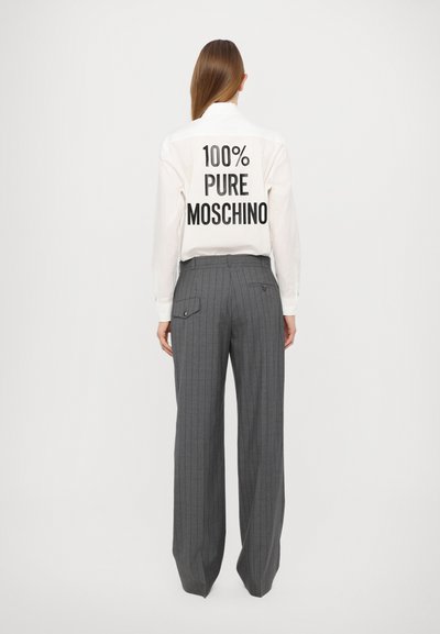 Valkoinen paita, jossa on musta teksti "100% PUHDAS MOSCHINO" selässä, yhdistettynä harmaisiin viivallisiin korkeavyötäröisiin housuihin, joissa on takataskut.