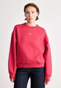 Sweatshirt en coton rouge avec un col rond, des poignets côtelés et une petite patch logo sur la poitrine, associé à un jean en denim bleu foncé.