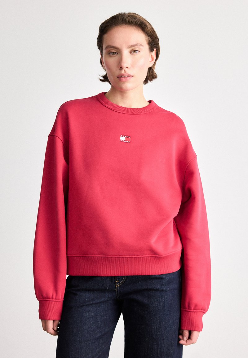 Sweatshirt en coton rouge avec un col rond, des poignets côtelés et une petite patch logo sur la poitrine, associé à un jean en denim bleu foncé.