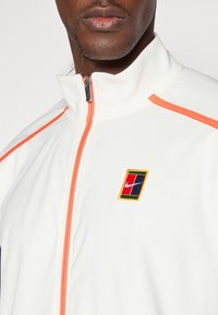 Λευκό φούτερ με φερμουάρ που διαθέτει πορτοκαλί λεπτομέρειες, κεντημένο λογότυπο Nike και υφασμάτινη υφή. Σημαντικά στοιχεία σχεδίασης περιλαμβάνουν ένα ψηλό γιακά.