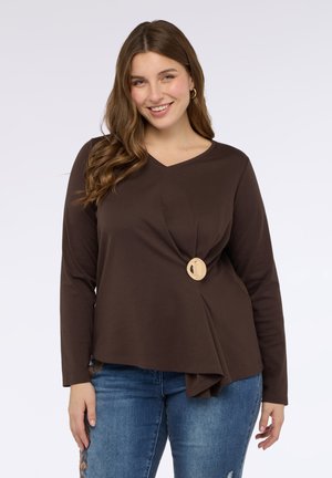 Maglietta marrone a maniche lunghe con scollo a V, caratterizzata da un design frontale torcato e un accento circolare in legno al centro. Abbinata a jeans blu.