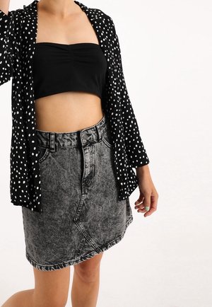 Kvinde iført en sort langærmet skjorte med prikker over en sort cropped top med en høj talje og acid wash denim nederdel.