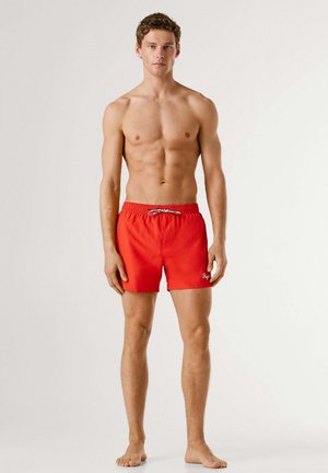 Noor särkideta mees paljajalu, kellel on seljas erksad punased ujumispüksid valge nööriga, seisab tavapärase heleda tausta ees.