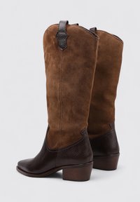 Bottes en daim marron jusqu'au genou avec un talon en cuir marron foncé texturé, une languette de traction en haut et un design courbé au niveau de la cheville.