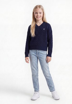 Junges Mädchen mit langen blonden Haaren, das einen dunkelblauen Pullover, hellblaue Jeans und weiße Turnschuhe trägt, steht und lächelt.