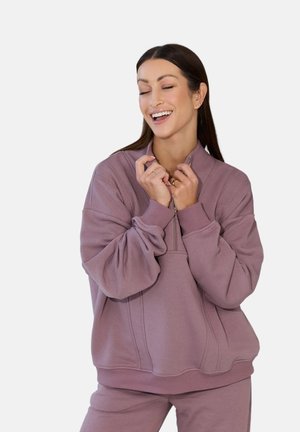 Uśmiechnięta kobieta z długimi włosami, ubrana w wrzosową bluzę i spodnie, trzymająca kołnierz bluzy obiema rękami na jednolitym tle.