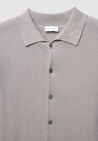 Helles graues Strick-Polohemd mit klassischem Kragen, fünf dunklen Knöpfen und strukturierter Oberfläche. Etikett mit der Aufschrift "CALLIOPE" am Kragen sichtbar.