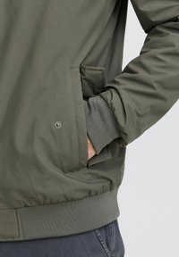 Maslinasto zeleni bomber s glatkom teksturom, rebrastim manžetama i bočnim džepom. Ima prilagođeni struk i minimalistički dizajn.