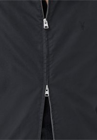 Zwarte ritsjack met een gladde buitenkant, voorzien van een zilveren rits en een subtiele geborduurde logo op de borst.